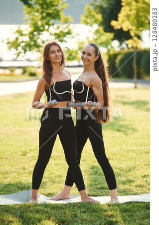 With dumbbells. Two women in sport clothes are doing exercises outdoors on the field With dumbbells. Two women in sport clothes are doing exercises outdoors on the field 128480183