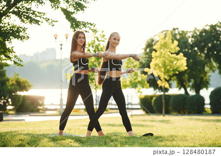 Dumbbells in hands. Two women in sport clothes are doing exercises outdoors on the field Dumbbells in hands. Two women in sport clothes are doing exercises outdoors on the field 128480187