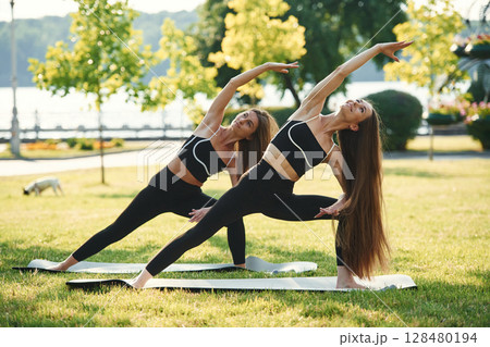 Asana named warrior in yoga. Two women in sport clothes are doing exercises outdoors on the field 128480194