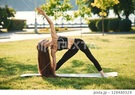 On the yoga mat. Beautiful young woman in sportive clothes is in the park at sunny daytime On the yoga mat. Beautiful young woman in sportive clothes is in the park at sunny daytime 128480329