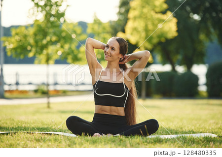 Sitting on the yoga mat. Beautiful young woman in sportive clothes is in the park at sunny daytime 128480335