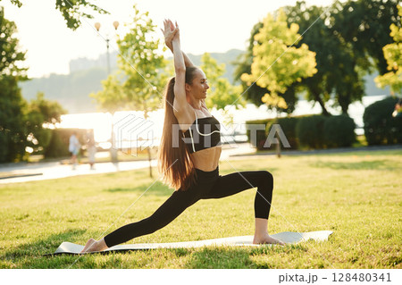 Doing squats. Beautiful young woman in sportive clothes is in the park at sunny daytime 128480341