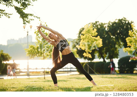 Standing, stretching the hands. Beautiful young woman in sportive clothes is in the park at sunny daytime Standing, stretching the hands. Beautiful young woman in sportive clothes is in the park at sunny daytime 128480353