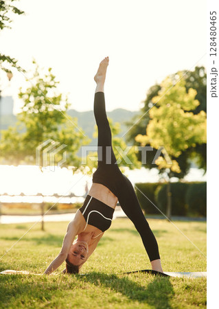 One leg is on the ground, another one is up. Beautiful young woman in sportive clothes is in the park at sunny daytime 128480465