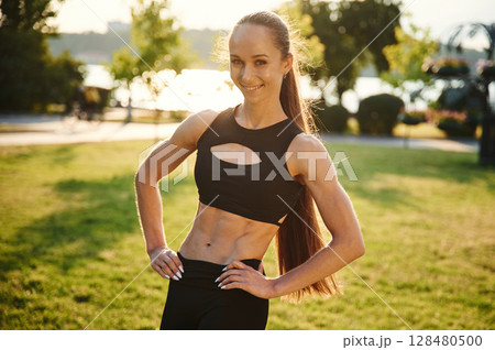 Showing abs. Beautiful young woman in sportive clothes is in the park at sunny daytime 128480500