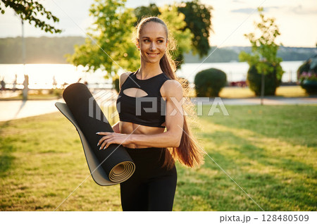 With yoga mat in hand. Beautiful young woman in sportive clothes is in the park at sunny daytime With yoga mat in hand. Beautiful young woman in sportive clothes is in the park at sunny daytime 128480509