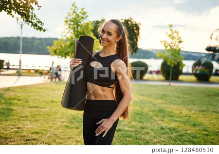 With yoga mat in hand. Beautiful young woman in sportive clothes is in the park at sunny daytime With yoga mat in hand. Beautiful young woman in sportive clothes is in the park at sunny daytime 128480510