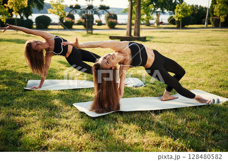 Warrior yoga asana, exercise. Two women in sport clothes are doing exercises outdoors on the field Warrior yoga asana, exercise. Two women in sport clothes are doing exercises outdoors on the field 128480582