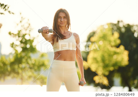 Holding dumbbell. Beautiful young woman in sportive clothes is in the park at sunny daytime 128480649