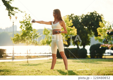 Holding dumbbell. Beautiful young woman in sportive clothes is in the park at sunny daytime 128480652