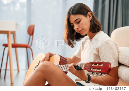 With classical guitar in hands. Beautiful woman in casual clothes is at home in domestic room 128482587