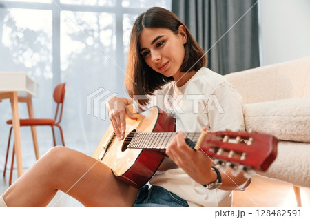 With classical guitar in hands. Beautiful woman in casual clothes is at home in domestic room With classical guitar in hands. Beautiful woman in casual clothes is at home in domestic room 128482591