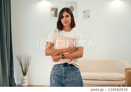 Standing and holding laptop in hands. Beautiful woman in casual clothes is at home in domestic room 128482616