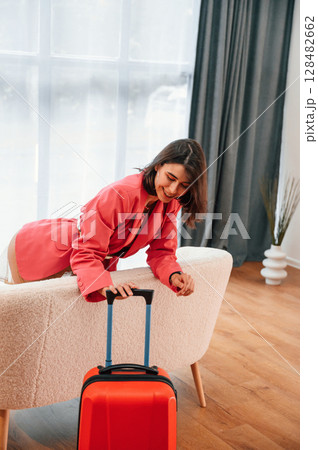 Looks at the suitcase. Woman in red clothes is indoors in the domestic room 128482662