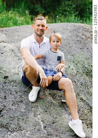 Smiling Father Sitting With Son On Large Rock In Nature 128482983