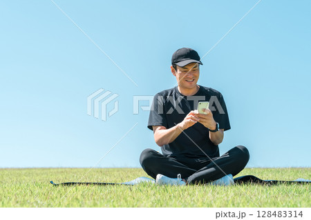 公園の芝生の上にヨガマットを引いて座りスマホを使うスポーツウェア姿のアジア人男性 公園の芝生の上にヨガマットを引いて座りスマホを使うスポーツウェア姿のアジア人男性 128483314