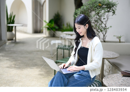 Smiling woman working on laptop at a stylish stone table surrounded by plants 128483934