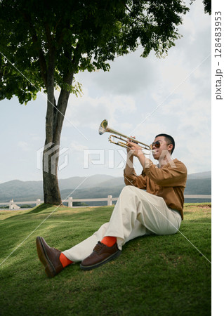 Young man sitting on grass under a tree, playing trumpet with mountains in the background 128483953