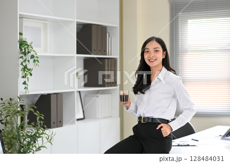 Confident businesswoman enjoying a coffee break while standing in a modern office space Confident businesswoman enjoying a coffee break while standing in a modern office space 128484031