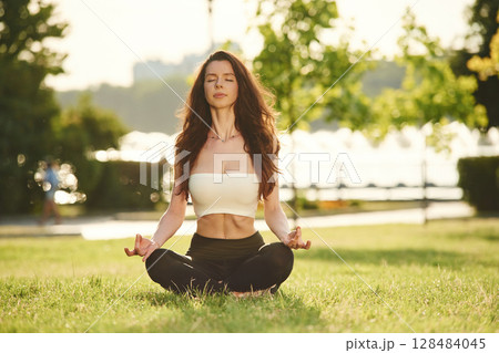 Meditating in lotus pose. Beautiful young woman in sportive clothes is in the park at sunny daytime 128484045
