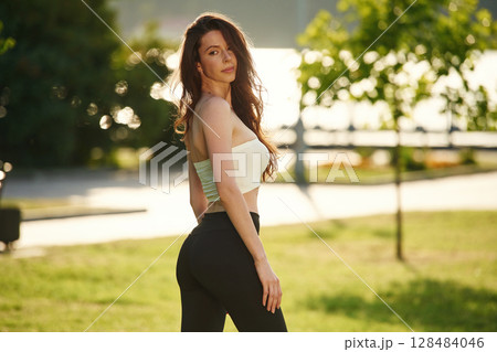 Standing, posing, showing body. Beautiful young woman in sportive clothes is in the park at sunny daytime Standing, posing, showing body. Beautiful young woman in sportive clothes is in the park at sunny daytime 128484046