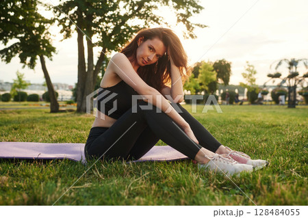 Sitting on the yoga mat against sunlight. Beautiful young woman in sportive clothes is in the park at sunny daytime Sitting on the yoga mat against sunlight. Beautiful young woman in sportive clothes is in the park at sunny daytime 128484055