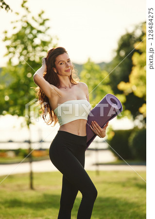 Holding yoga mat in hands. Beautiful young woman in sportive clothes is in the park at sunny daytime Holding yoga mat in hands. Beautiful young woman in sportive clothes is in the park at sunny daytime 128484275