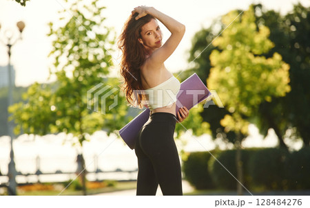 Holding yoga mat in hands. Beautiful young woman in sportive clothes is in the park at sunny daytime 128484276