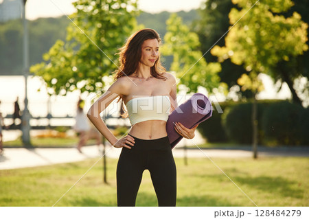 Purple colored yoga mat in hands. Beautiful young woman in sportive clothes is in the park at sunny daytime 128484279