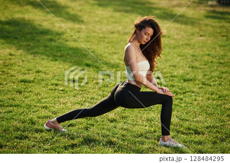 On green grass, stretching the legs. Beautiful young woman in sportive clothes is in the park at sunny daytime On green grass, stretching the legs. Beautiful young woman in sportive clothes is in the park at sunny daytime 128484295