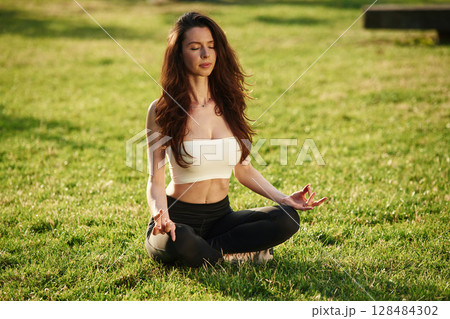 Meditating in lotus pose. Beautiful young woman in sportive clothes is in the park at sunny daytime Meditating in lotus pose. Beautiful young woman in sportive clothes is in the park at sunny daytime 128484302