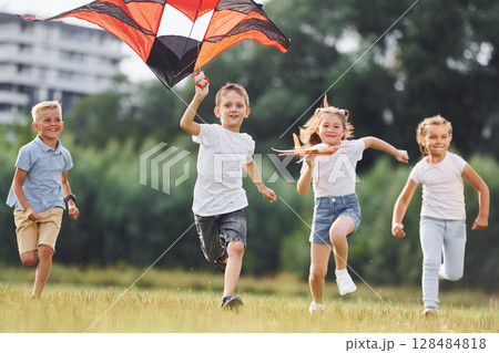 Happy children. Group of kids are running and playing with kite on green field 128484818