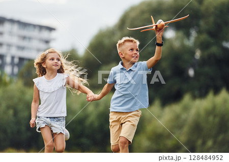 Running forward. Boy and girl are playing with toy plane on the green field Running forward. Boy and girl are playing with toy plane on the green field 128484952