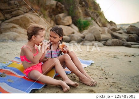 Joyful sisters enjoying snacks on the sandy beach at sunset 128485003
