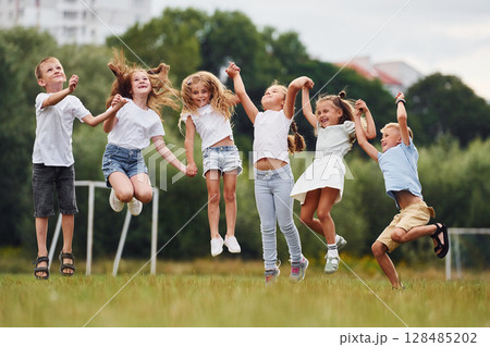 Holding each other by hands and jumping. Kids are having fun on the field at daytime together Holding each other by hands and jumping. Kids are having fun on the field at daytime together 128485202