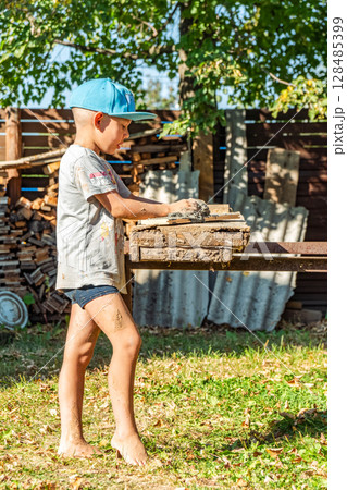 Young boy in a cap cheerfully creates with clay, standing barefoot and covered in dirt, near a rustic wooden yard table Young boy in a cap cheerfully creates with clay, standing barefoot and covered in dirt, near a rustic wooden yard table 128485399