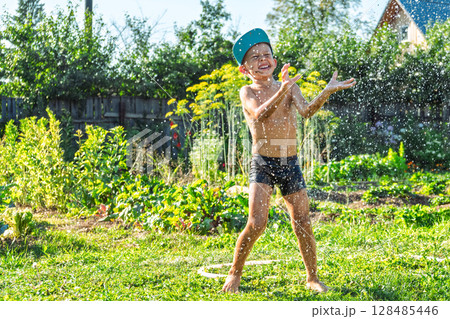 Joyful boy in a cap enjoys splashing water from a hose, playfully cleaning himself in a vibrant garden under bright sunshine Joyful boy in a cap enjoys splashing water from a hose, playfully cleaning himself in a vibrant garden under bright sunshine 128485446
