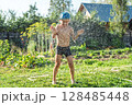 Joyful boy in a cap enjoys splashing water from a hose, playfully cleaning himself in a vibrant garden under bright sunshine 128485448