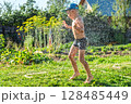 Joyful boy in a cap enjoys splashing water from a hose, playfully cleaning himself in a vibrant garden under bright sunshine 128485449