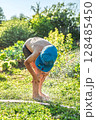 Joyful boy in a cap enjoys splashing water from a hose, playfully cleaning himself in a vibrant garden under bright sunshine 128485450