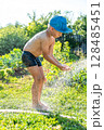 Joyful boy in a cap enjoys splashing water from a hose, playfully cleaning himself in a vibrant garden under bright sunshine 128485451
