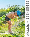 Joyful boy in a cap enjoys splashing water from a hose, playfully cleaning himself in a vibrant garden under bright sunshine 128485452