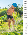 Joyful boy in a cap enjoys splashing water from a hose, playfully cleaning himself in a vibrant garden under bright sunshine 128485453
