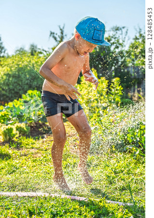 Joyful boy in a cap enjoys splashing water from a hose, playfully cleaning himself in a vibrant garden under bright sunshine 128485453