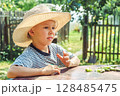Small boy in a straw hat focuses on shelling beans while sitting at a table outside, surrounded by greenery 128485475
