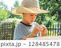Small boy in a straw hat focuses on shelling beans while sitting at a table outside, surrounded by greenery 128485478