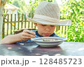 Young boy wearing a hat savours a bowl of soup, sitting comfortably at a rustic wooden table in a sunny garden 128485723