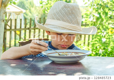 Young boy wearing a hat savours a bowl of soup, sitting comfortably at a rustic wooden table in a sunny garden 128485723
