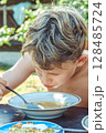 Boy with thick hair enjoys lunch at a wooden table in a sunny country garden setting 128485724
