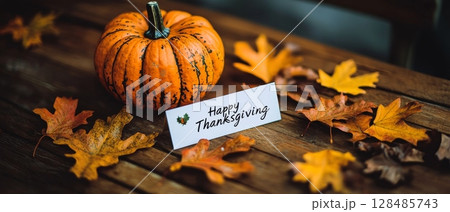A rustic wooden table adorned with a striped pumpkin, colorful autumn leaves, and a 'Happy Thanksgiving' card, creating a warm and inviting holiday scene 128485743